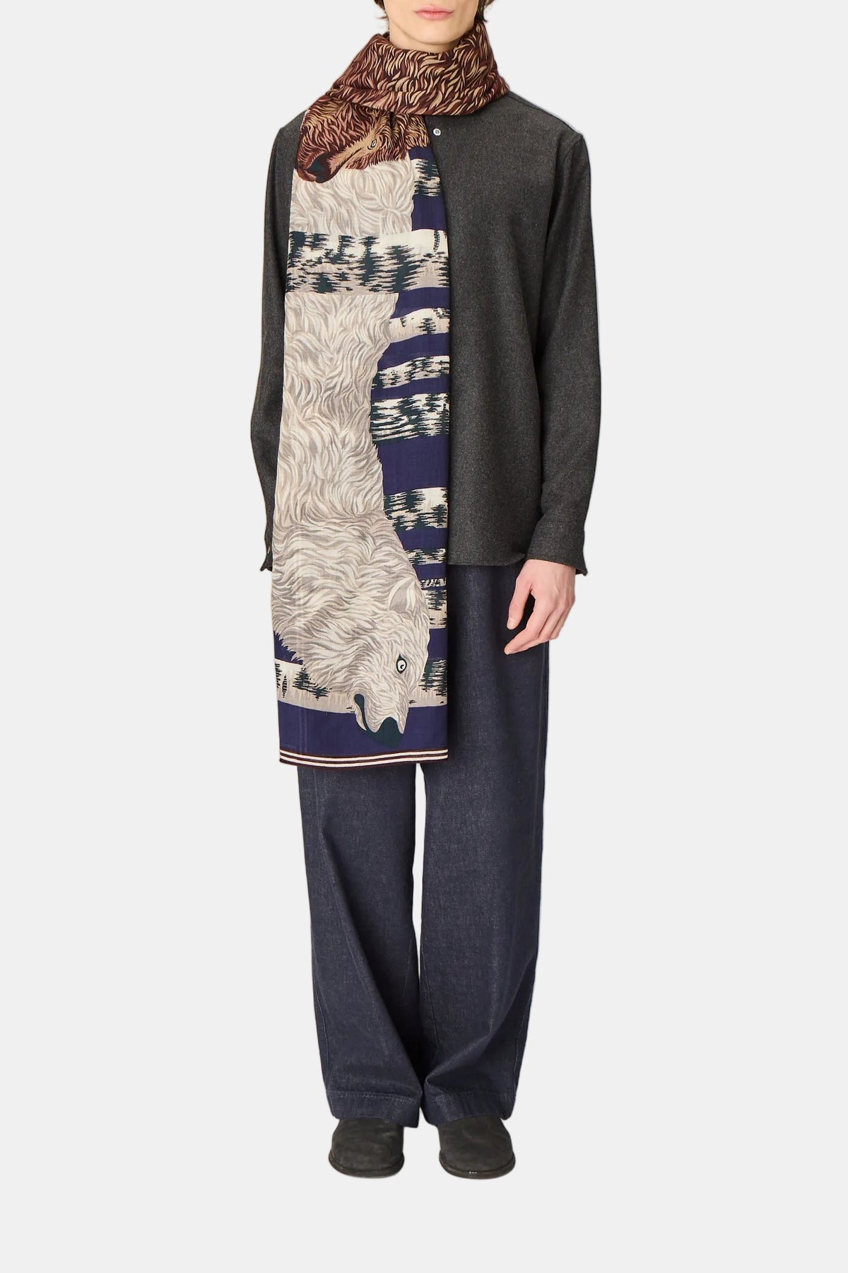 A model stands against a light gray studio background wearing a dark charcoal cardigan, navy wide-leg trousers, a brown patterned scarf, and a long printed scarf featuring a wolf and blue stripes.