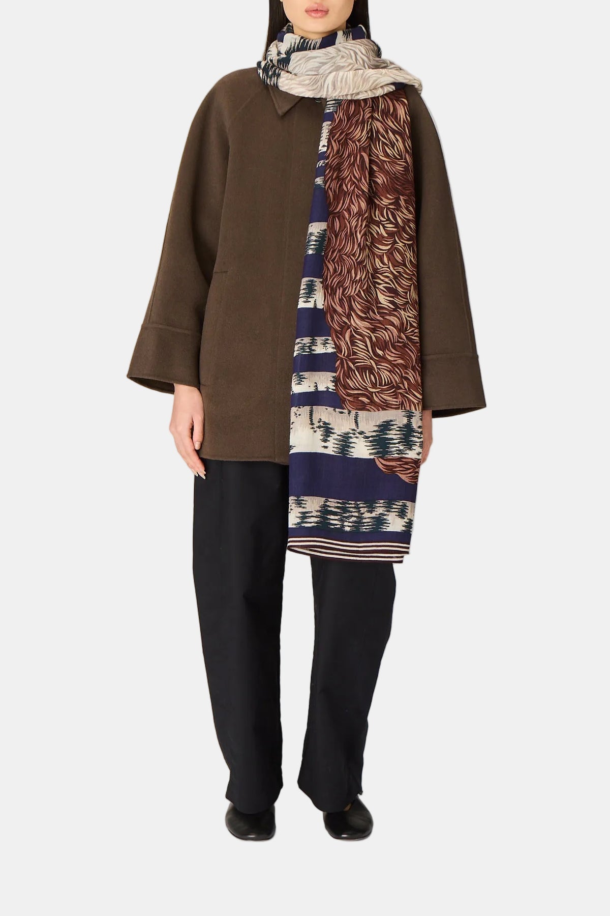 A model stands against a light gray studio backdrop wearing a brown coat, black wide-leg pants, and a long patterned scarf with blue, brown, and cream sections.