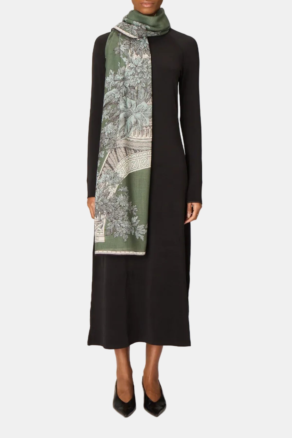 A model stands against a light gray studio backdrop wearing a long-sleeve black midi dress and black pointed shoes, with a green and white patterned scarf wrapped around the neck.