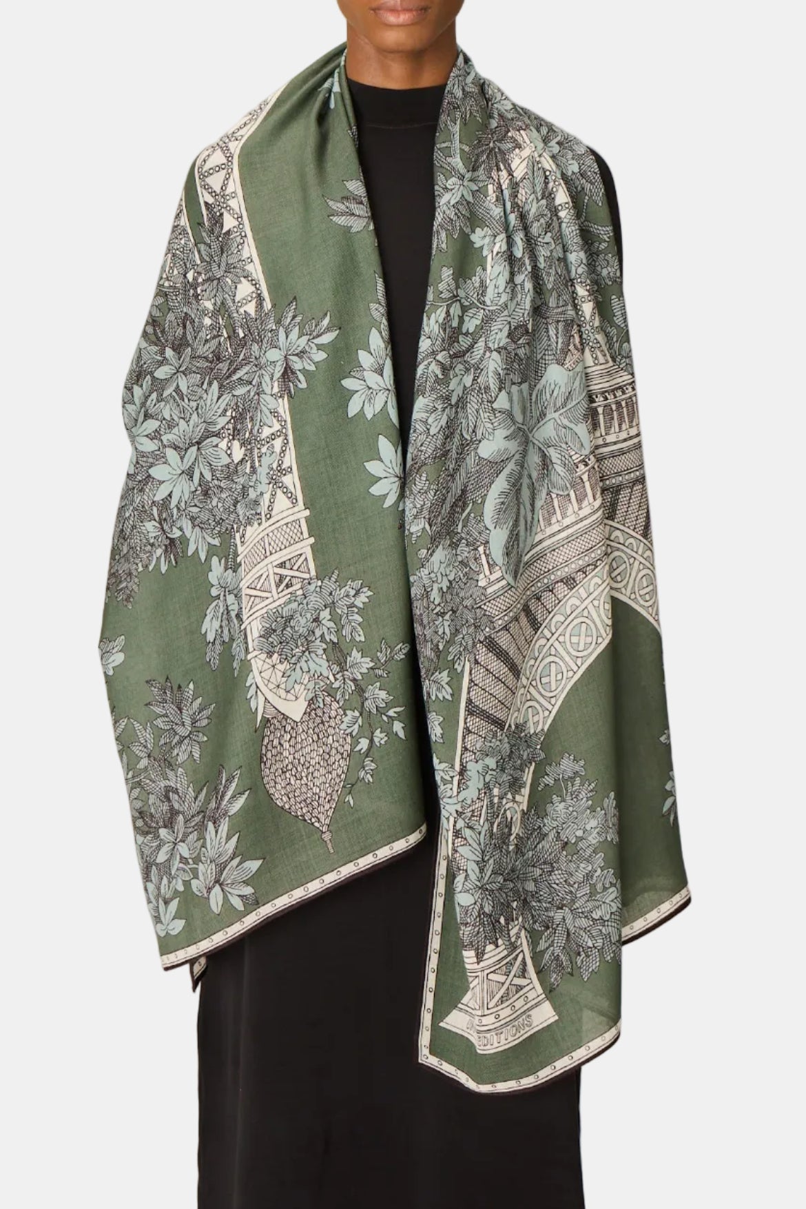 A model wearing a sage green silk scarf with a detailed botanical and architectural print, draped over their shoulders and paired with a black top and black trousers, set against a plain light gray studio background.