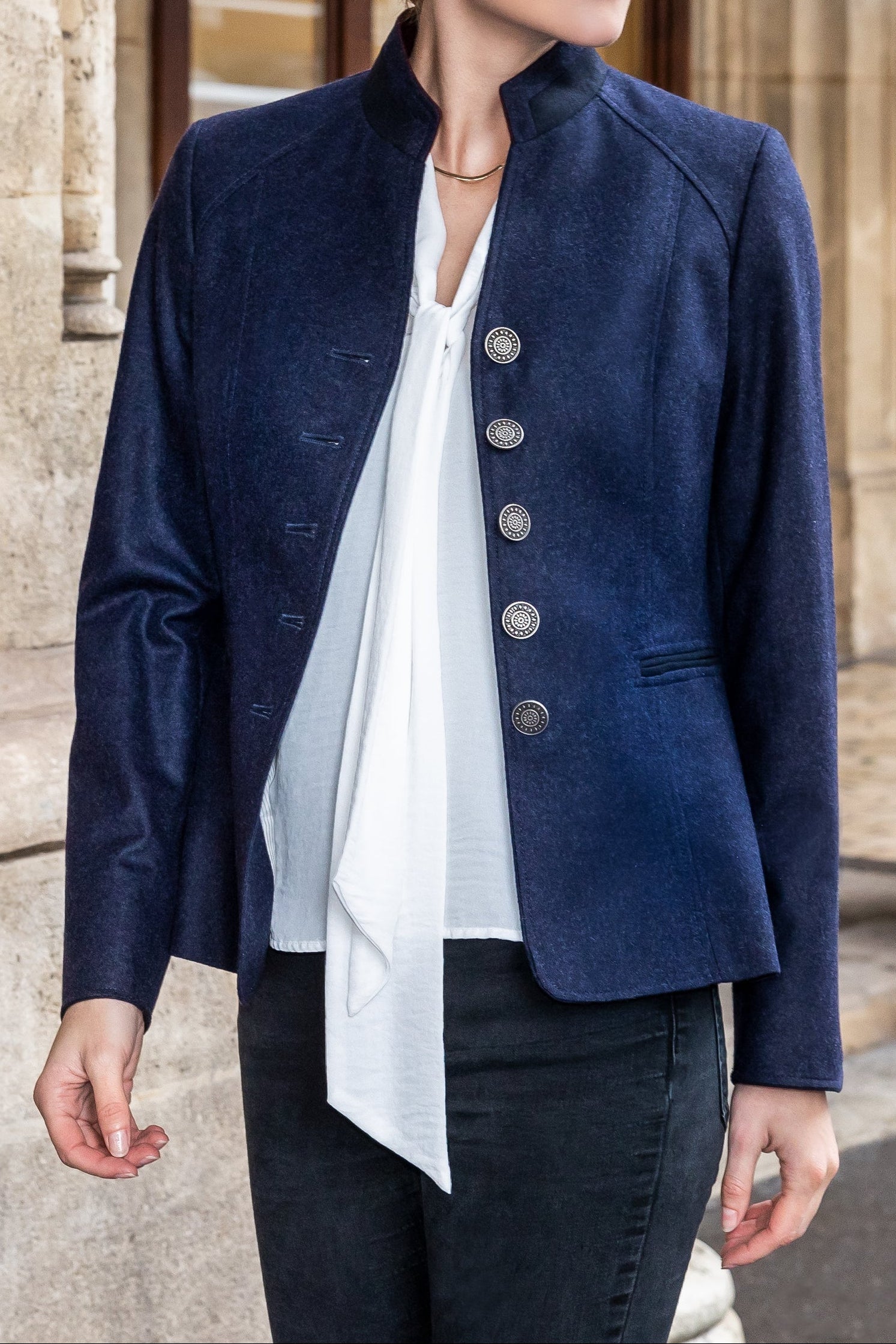 Woman wearing a navy blue jacket with a white blouse and black pants, standing in front of a stone building.