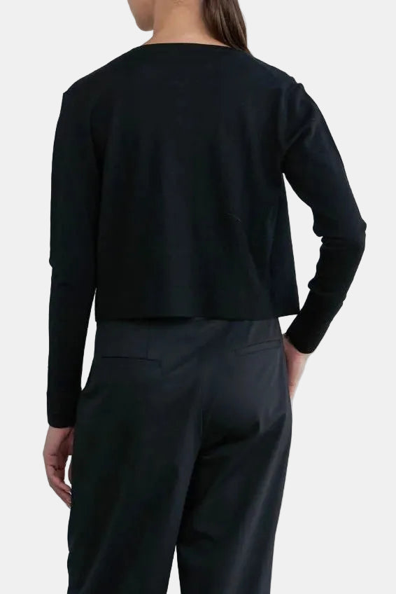 A model shown from the back wearing a black long-sleeve cropped top and black wide-leg trousers, standing against a minimalist light gray studio background.