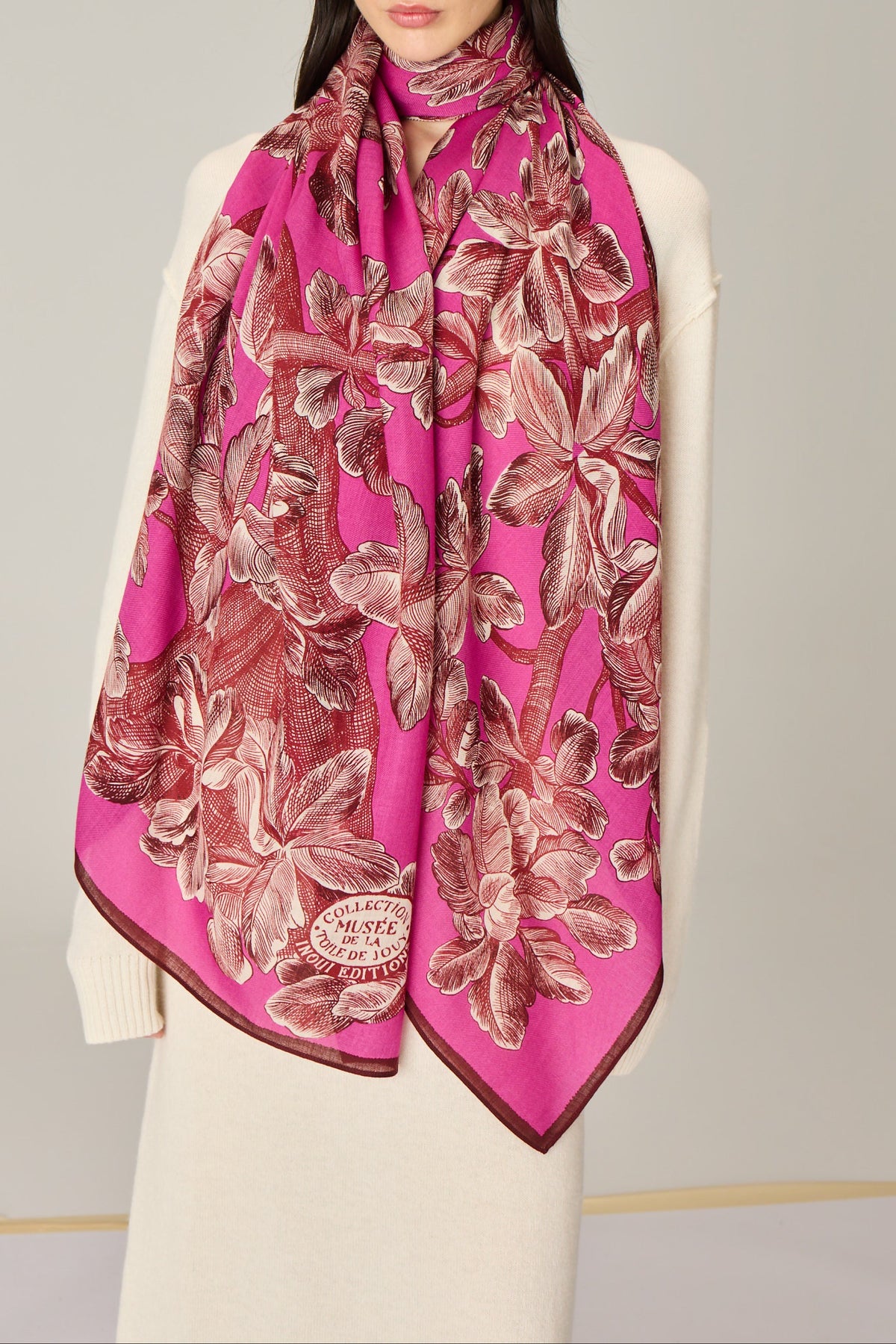 A woman wearing a bright pink silk scarf with a large floral print in dusty rose and brown, styled over a cream-colored sweater.
