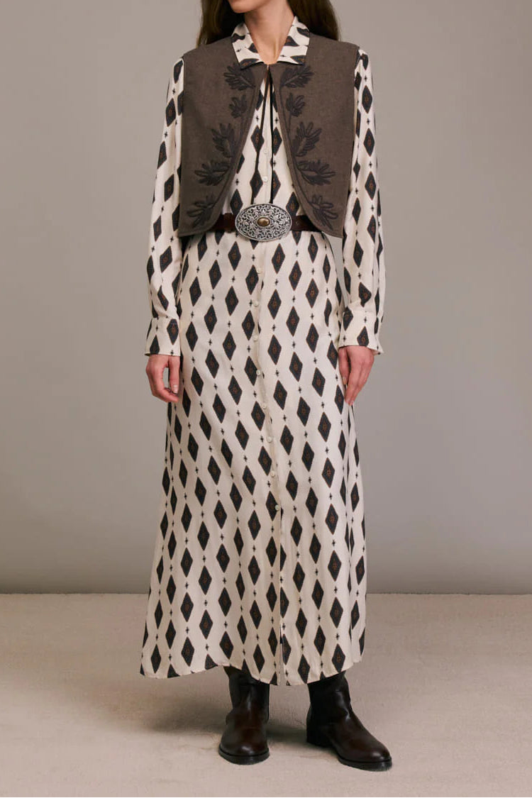 Woman wearing a long patterned dress with a vest against a plain background