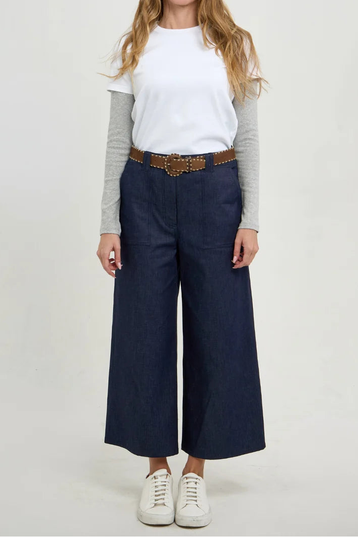 A woman modeling a pair of dark blue wide-leg cropped pants with a brown belt, styled with a white long-sleeve tee, a white t-shirt, and white sneakers against a light gray studio background.