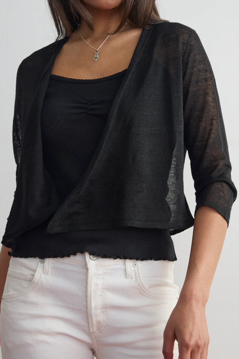 A woman wearing a black sheer open-front shrug or cardigan over a black top and white pants, shown from the neck to the hips against a plain light gray studio background.