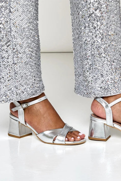 Silver metallic block-heel sandals with ankle straps worn with silver sequined pants, shown on a model's feet against a clean white background.