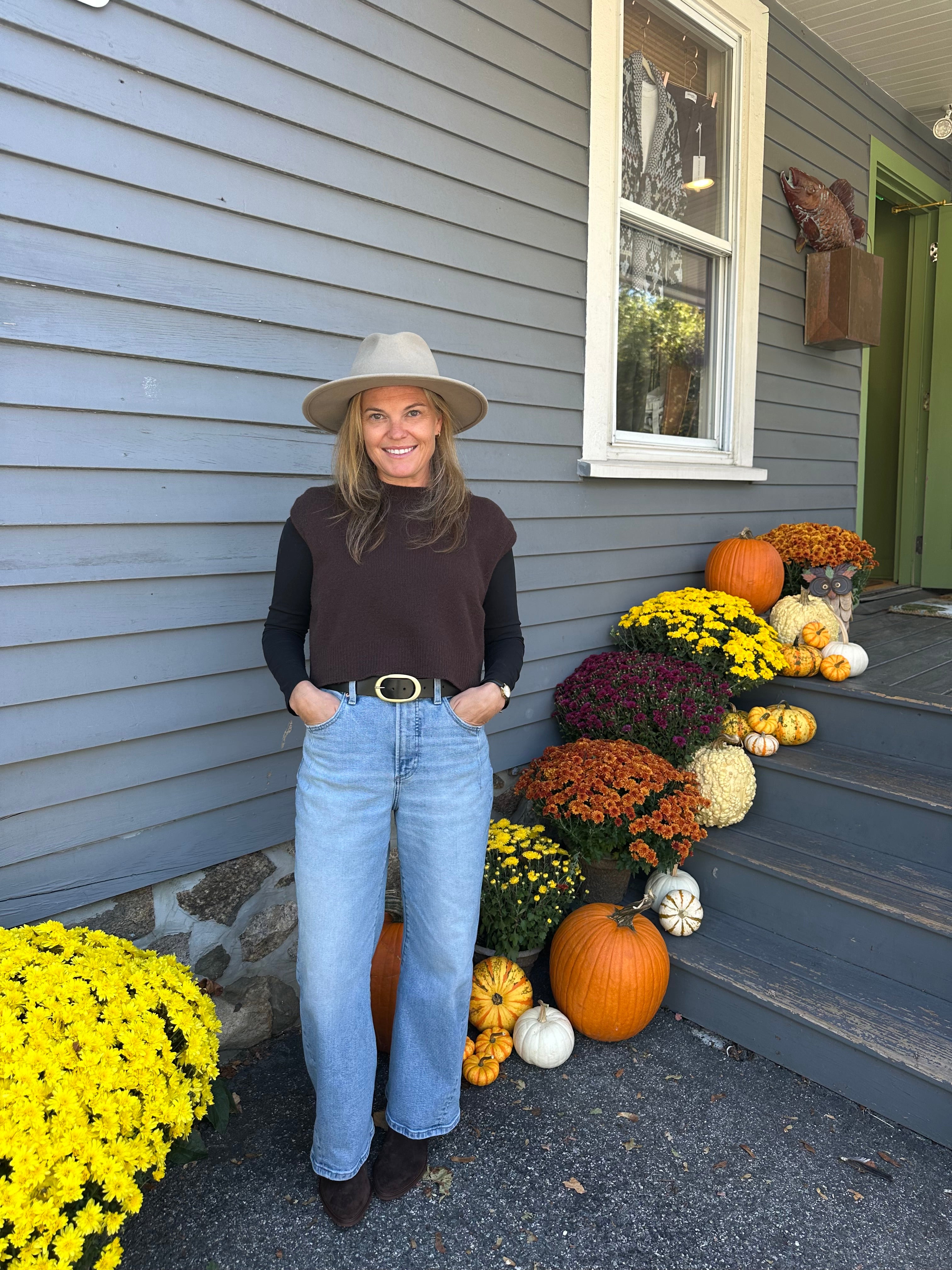 Person standing on a porch with autumn decorations including pumpkins and flowers.