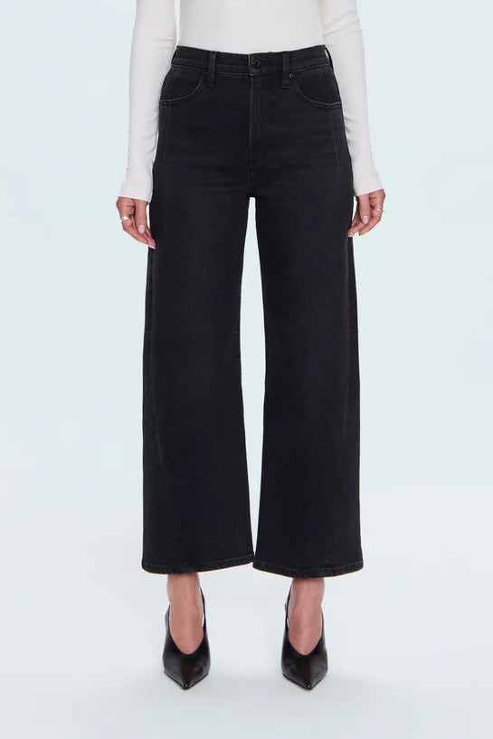 A model stands against a light gray studio background wearing a fitted white long-sleeve top, black wide-leg cropped pants, and black pointed heels.