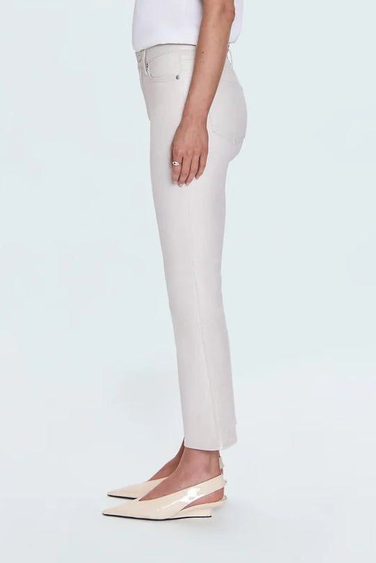 A woman in profile wearing a white short-sleeve top, light beige cropped trousers, and beige slingback wedge flats, standing against a very light gray studio background.