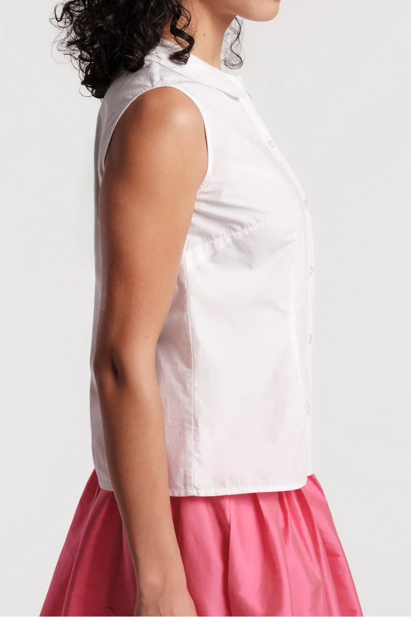 A woman shown in side profile wearing a sleeveless white high-neck top and a pink skirt against a light gray studio background.
