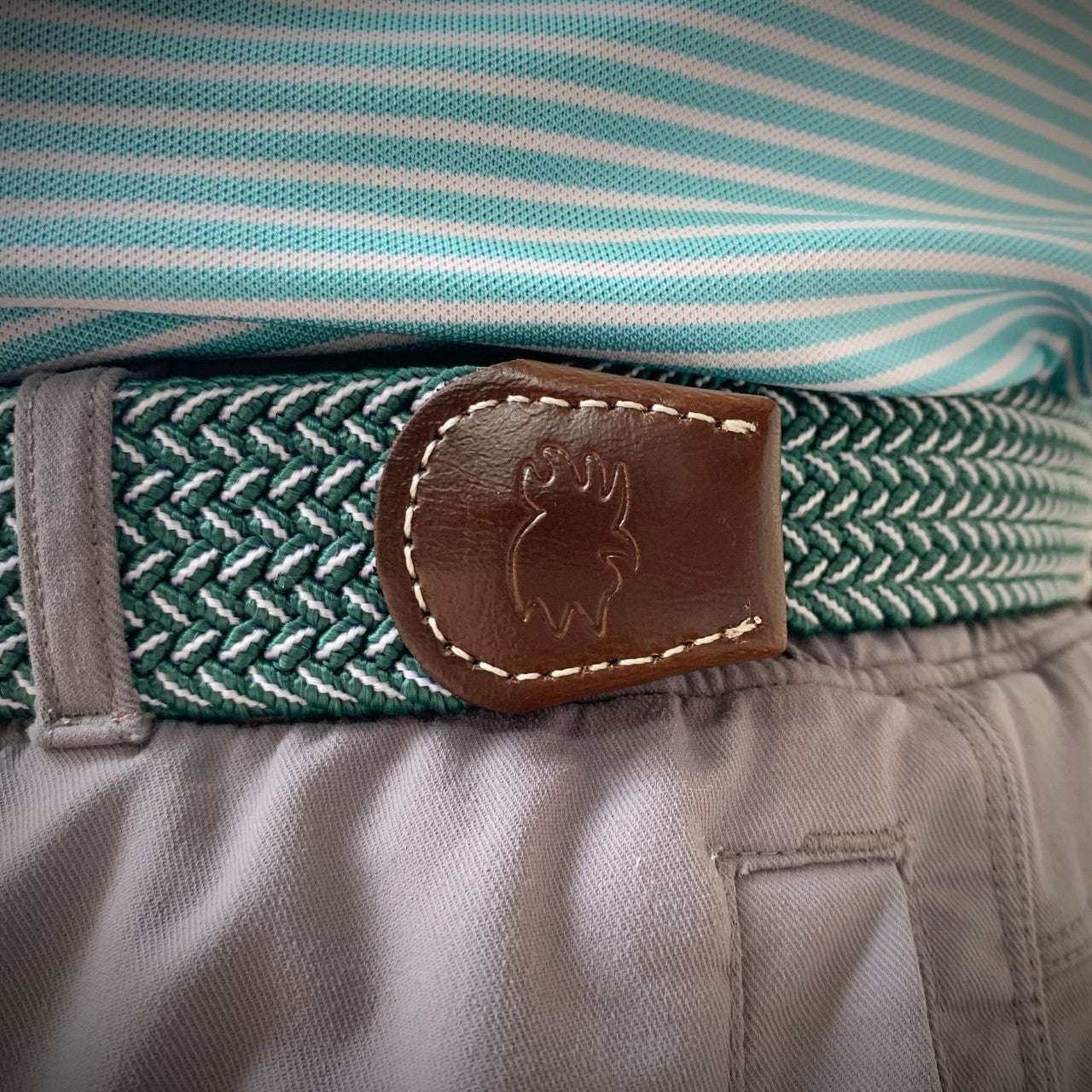 Close-up of a woven belt with a brown leather logo patch, worn around the waist over a striped shirt and gray pants.