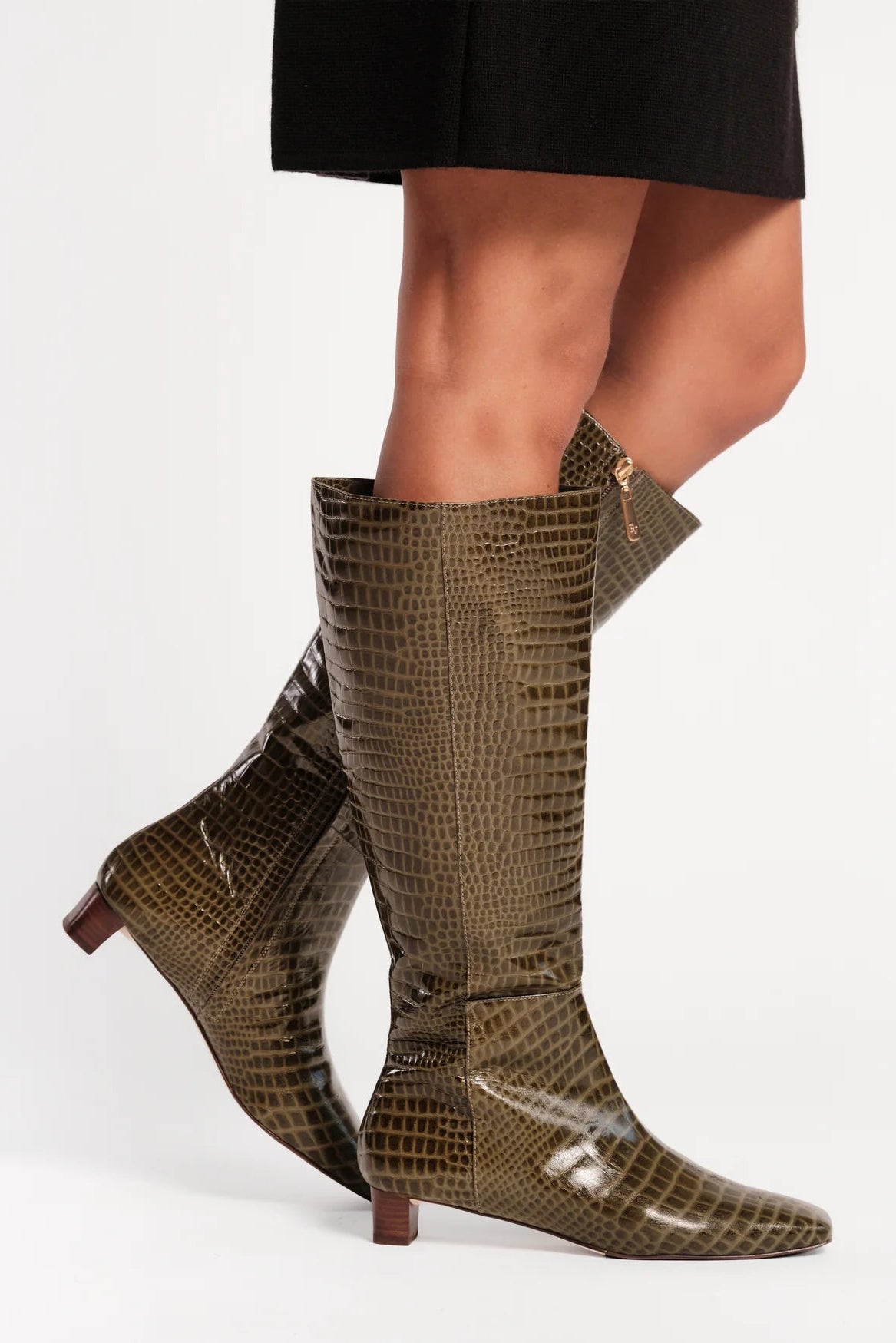 A pair of knee-high pointed-toe boots with a glossy crocodile-embossed pattern in olive brown and black, shown on a model's legs against a plain white studio background.