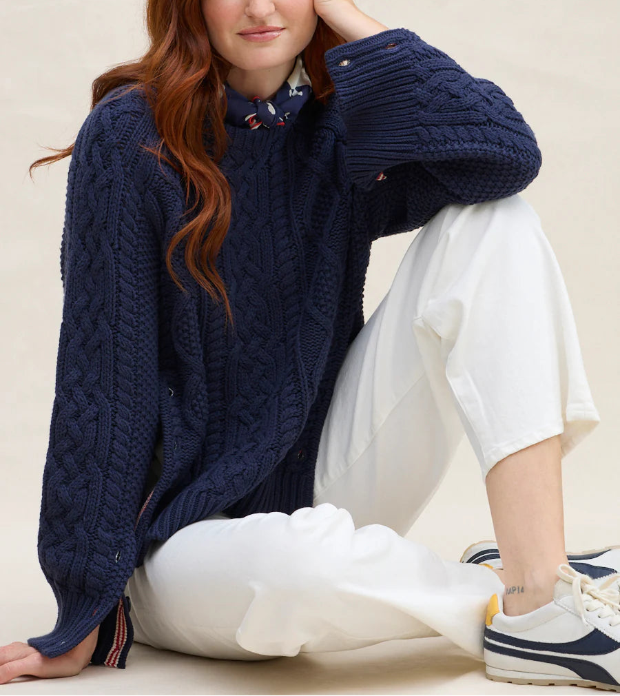 Woman wearing a navy blue cable knit sweater with a white outfit underneath, sitting on a beige surface.