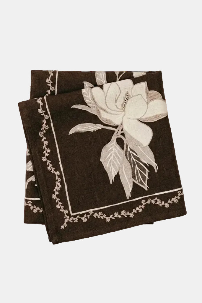 A folded dark brown linen napkin featuring a large cream-colored magnolia flower and delicate floral border details, set against a plain white background.