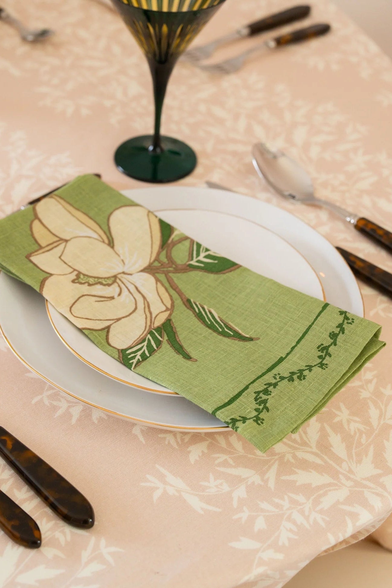 A green linen napkin with a large cream magnolia flower and leaf pattern, folded on a white plate at a table setting. The table is covered with a light peach floral tablecloth, surrounded by silver cutlery with dark tortoiseshell handles and a dark green stemmed glass.