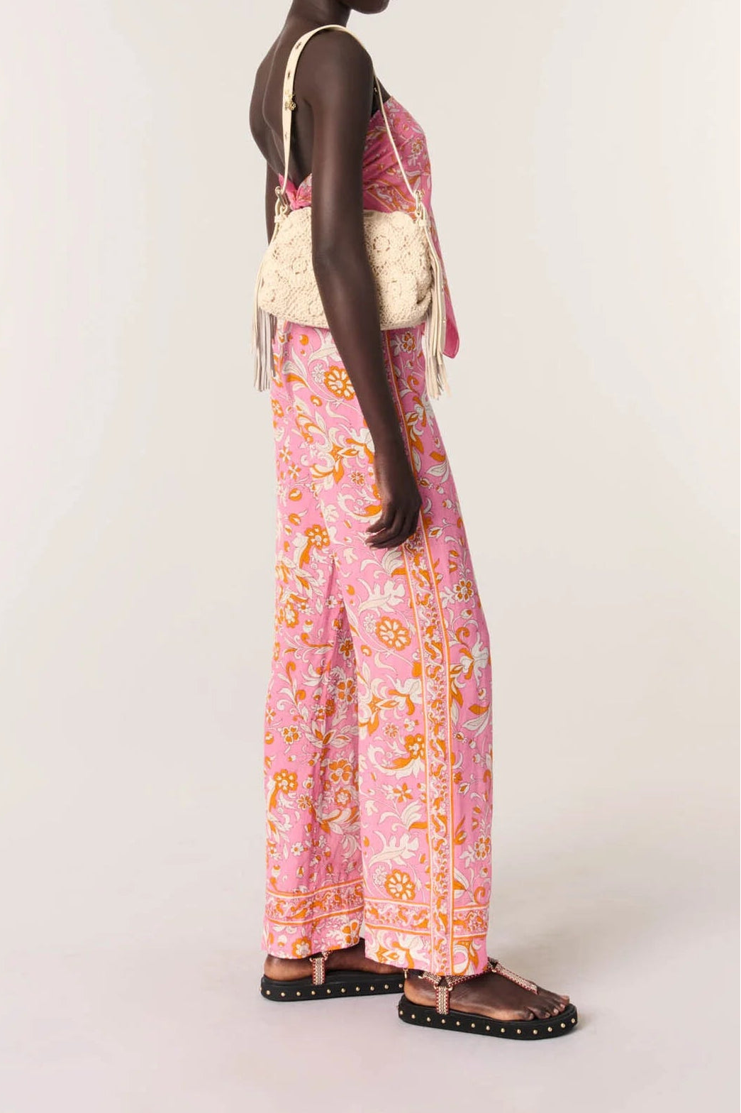 A model is shown from the back wearing a pink and orange patterned halter jumpsuit with wide-leg pants, a cream woven shoulder bag, and black studded sandals, set against a plain light gray studio background.