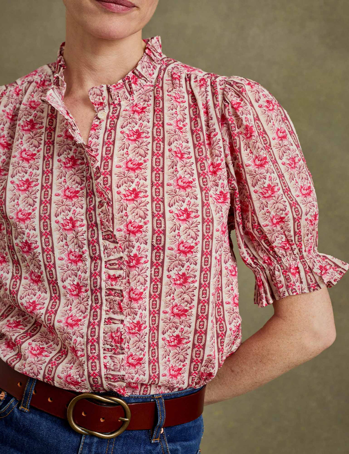 A close-up of a woman wearing a short puff-sleeve blouse with a vertical toile-style print in pink and red on a light beige background, paired with a brown belt and blue jeans against a neutral textured backdrop.