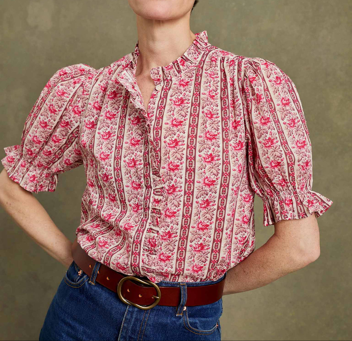 A woman wearing a pink and red patterned short-sleeve blouse with puff sleeves and a ruffled collar, paired with blue jeans and a brown belt, against a textured olive-green background.
