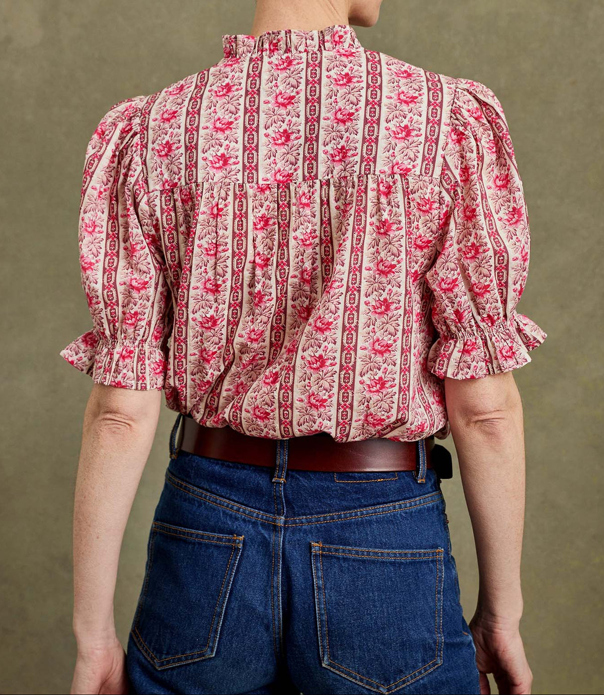 A person shown from the back wearing a pink and red floral puff-sleeve blouse with blue jeans against a textured olive-brown studio background.