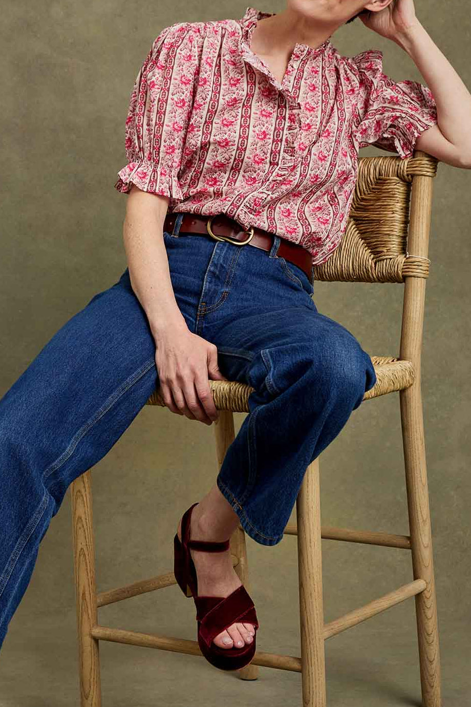 A model sits on a wooden stool wearing a pink and white patterned short-sleeve blouse, blue wide-leg jeans, and burgundy platform sandals against a textured olive-green studio backdrop.