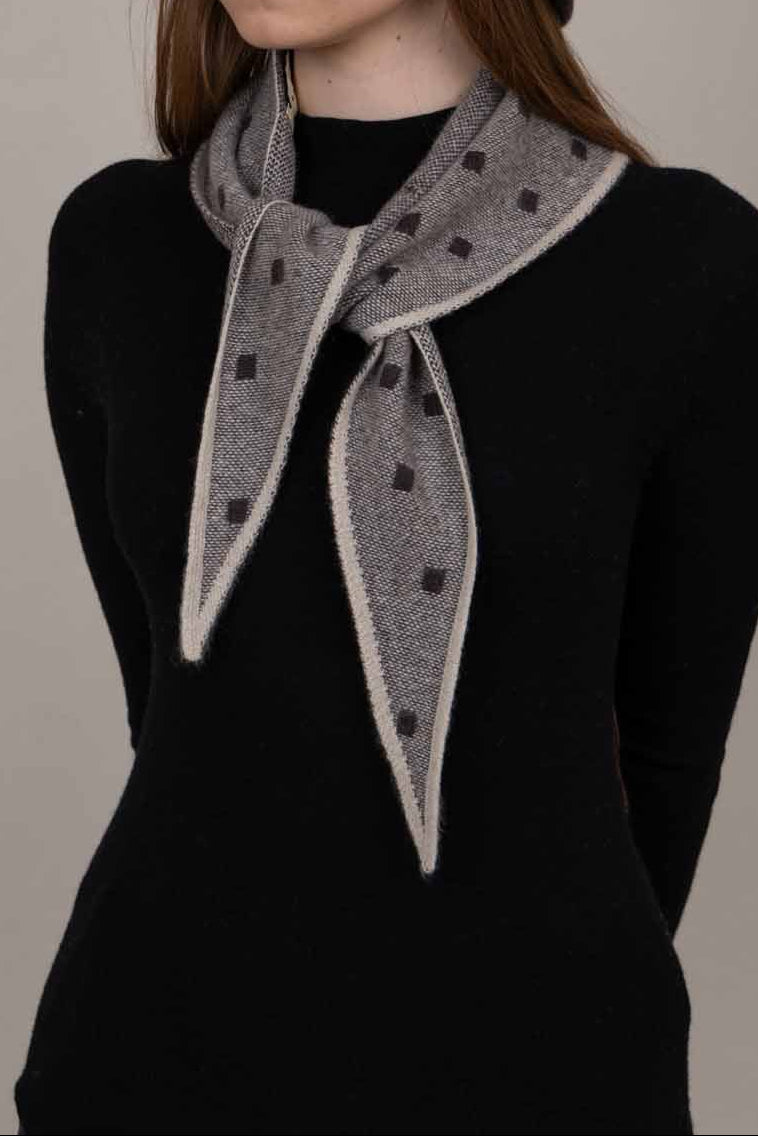 A woman wearing a dark brown felt cloche hat and a gray patterned scarf with white trim, styled with a black long-sleeve top against a plain beige background.