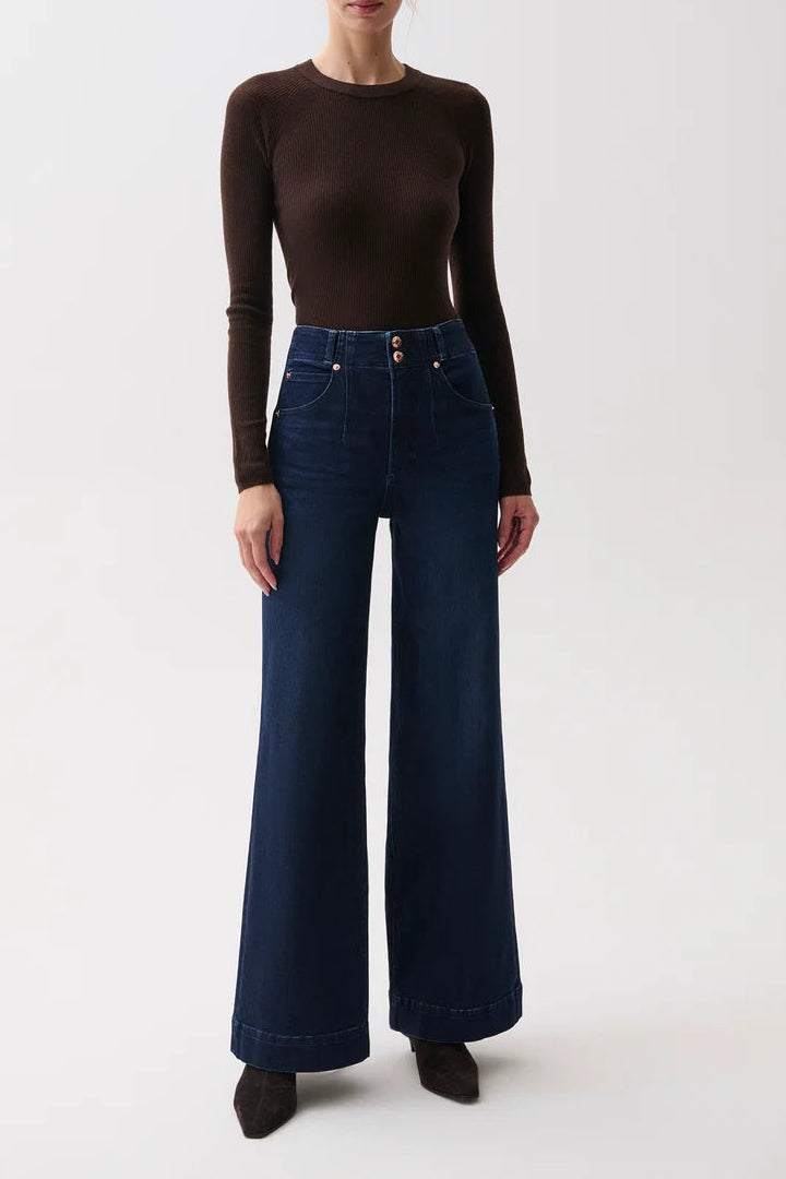 A woman stands against a plain light gray studio background wearing a fitted dark brown long-sleeve top and dark blue wide-leg jeans with dark pointed shoes.