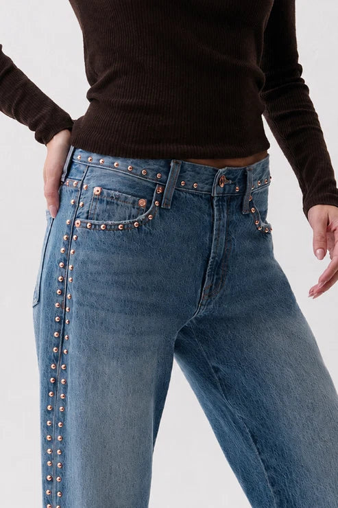 Close-up of a person wearing blue jeans with copper-toned studs along the pocket edges and waistband, paired with a dark brown ribbed long-sleeve top against a light gray studio background.