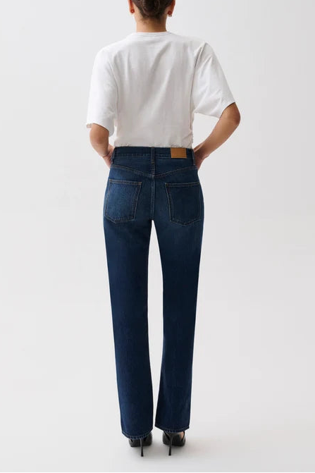A model viewed from behind wearing dark blue straight-leg jeans, a white t-shirt, and black heels against a plain white studio background.