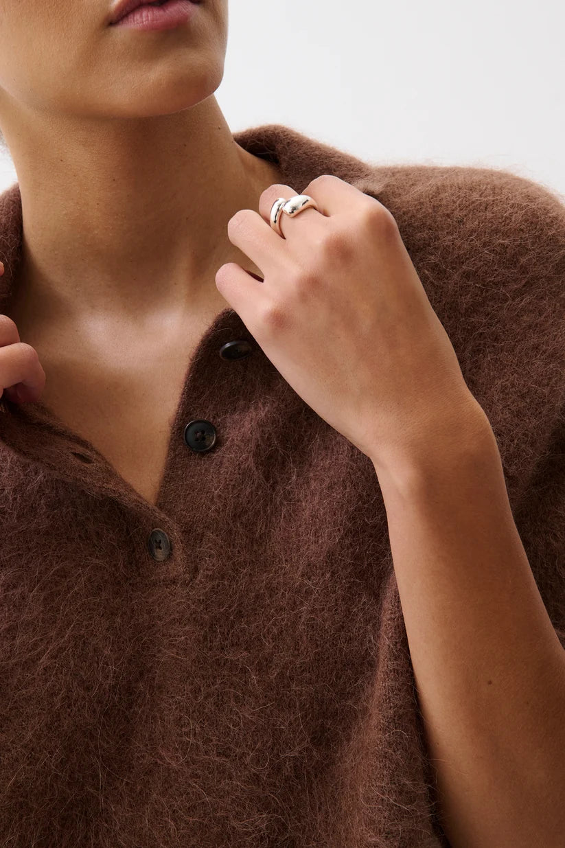 Close-up of a person wearing a fuzzy brown knit top and silver rings on their fingers, against a plain light background.