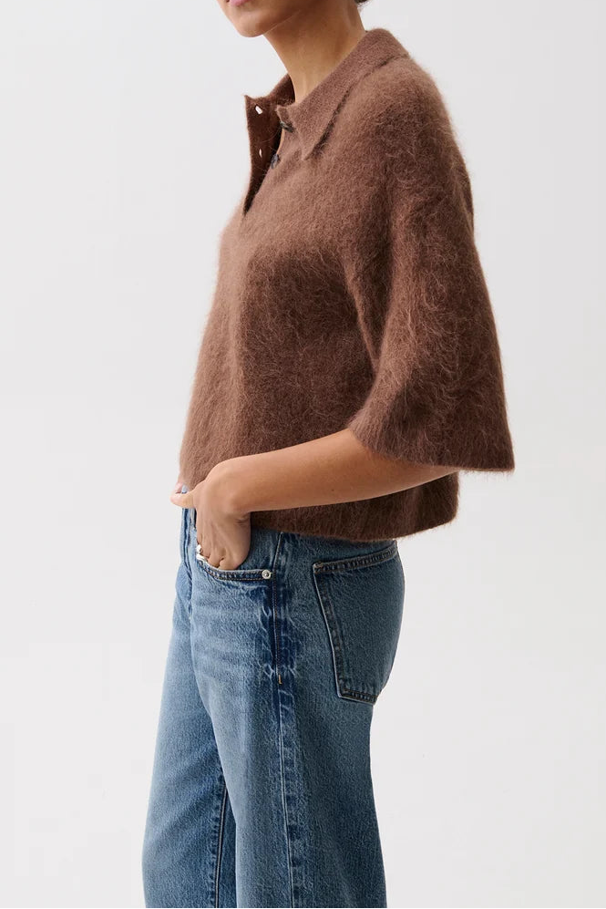 A woman modeling a brown fuzzy short-sleeve polo shirt with blue jeans against a plain light gray studio background.