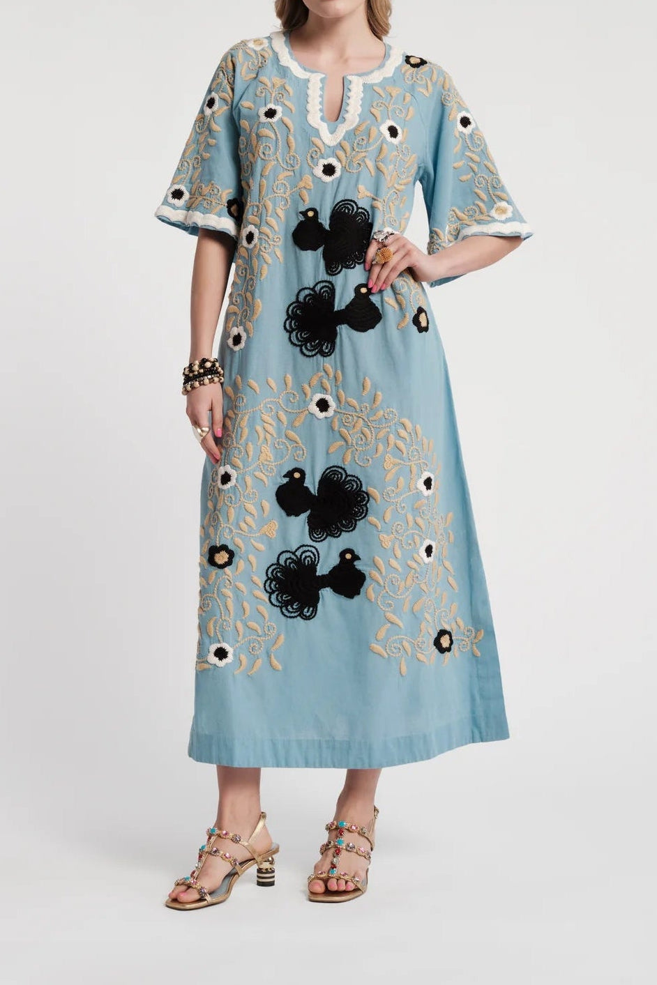 A woman modeling a light blue kaftan dress with gold embroidery, black floral appliqués, and white lace trim at the neckline, photographed against a plain light gray studio background.