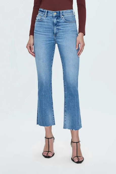 A woman modeling a dark brown long-sleeve crewneck top and light blue cropped flare jeans with black strappy heels, standing against a light gray studio background.