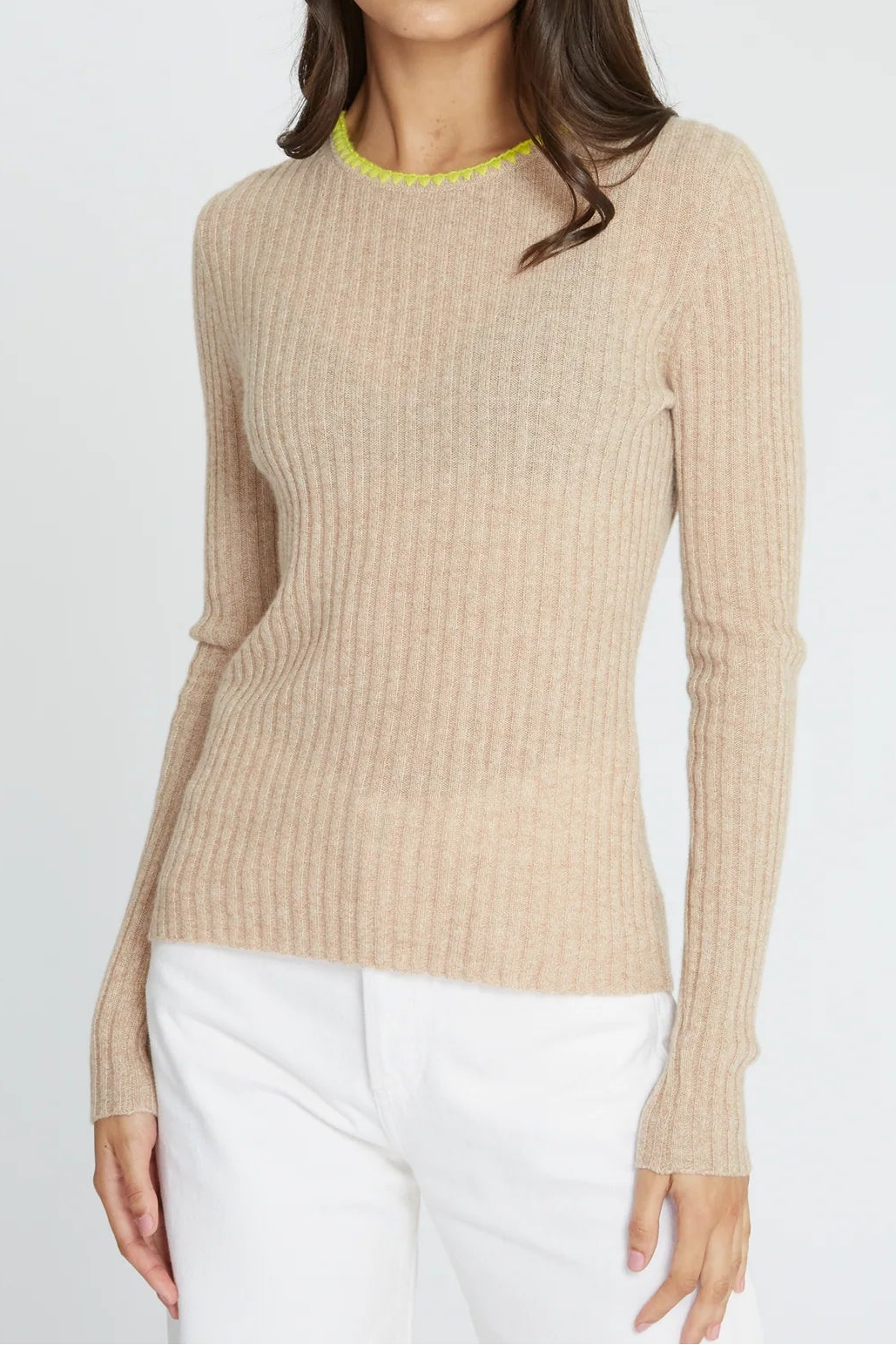 A woman modeling a beige ribbed knit crewneck sweater with a bright yellow-green collar trim, paired with white pants, against a light gray studio background.