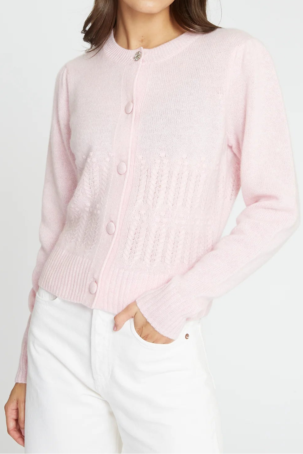 A woman wearing a light pink knit cardigan with button-front closure and white pants, photographed against a plain light gray studio background.