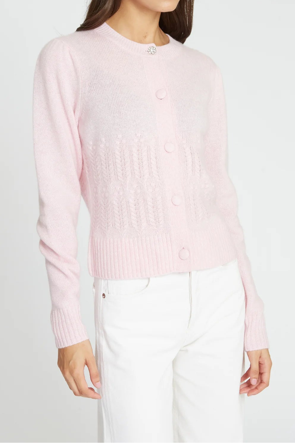 A woman modeling a light pink knit cardigan sweater with white pants against a plain light gray studio background.