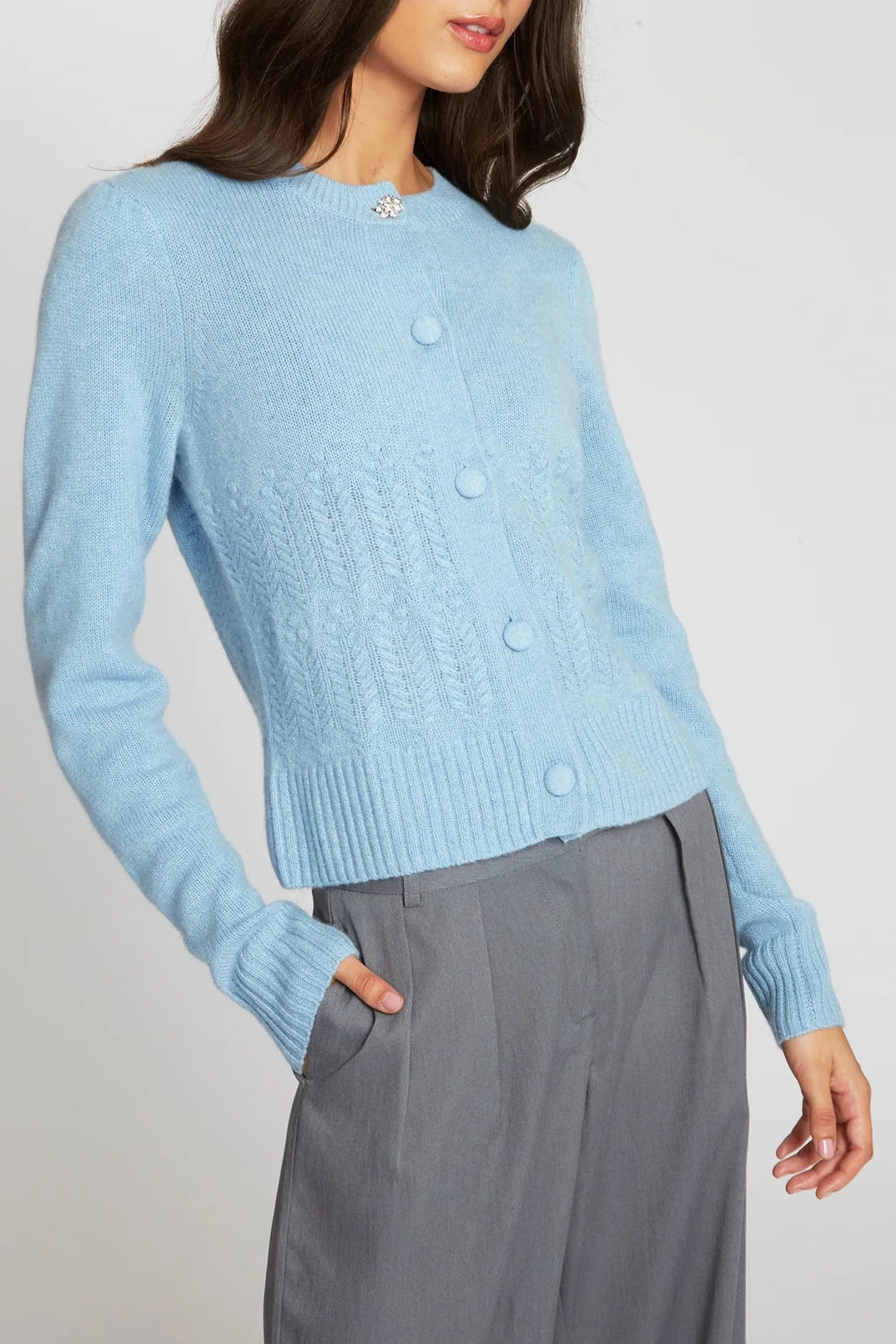 A woman wearing a light blue knit cardigan with buttons and gray trousers, photographed against a plain light gray studio background.