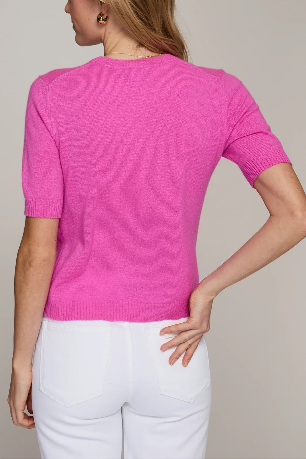 Woman wearing a bright pink sweater and white pants against a neutral background