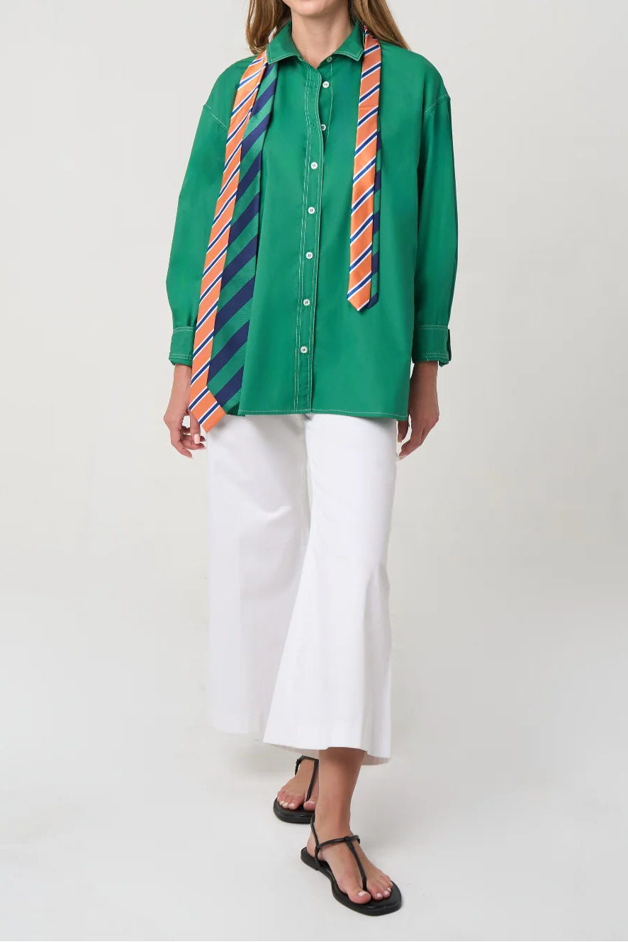 A woman modeling a bright green button-up shirt with two striped neckties draped over the collar, paired with white wide-leg cropped pants and black sandals, against a light gray studio background.