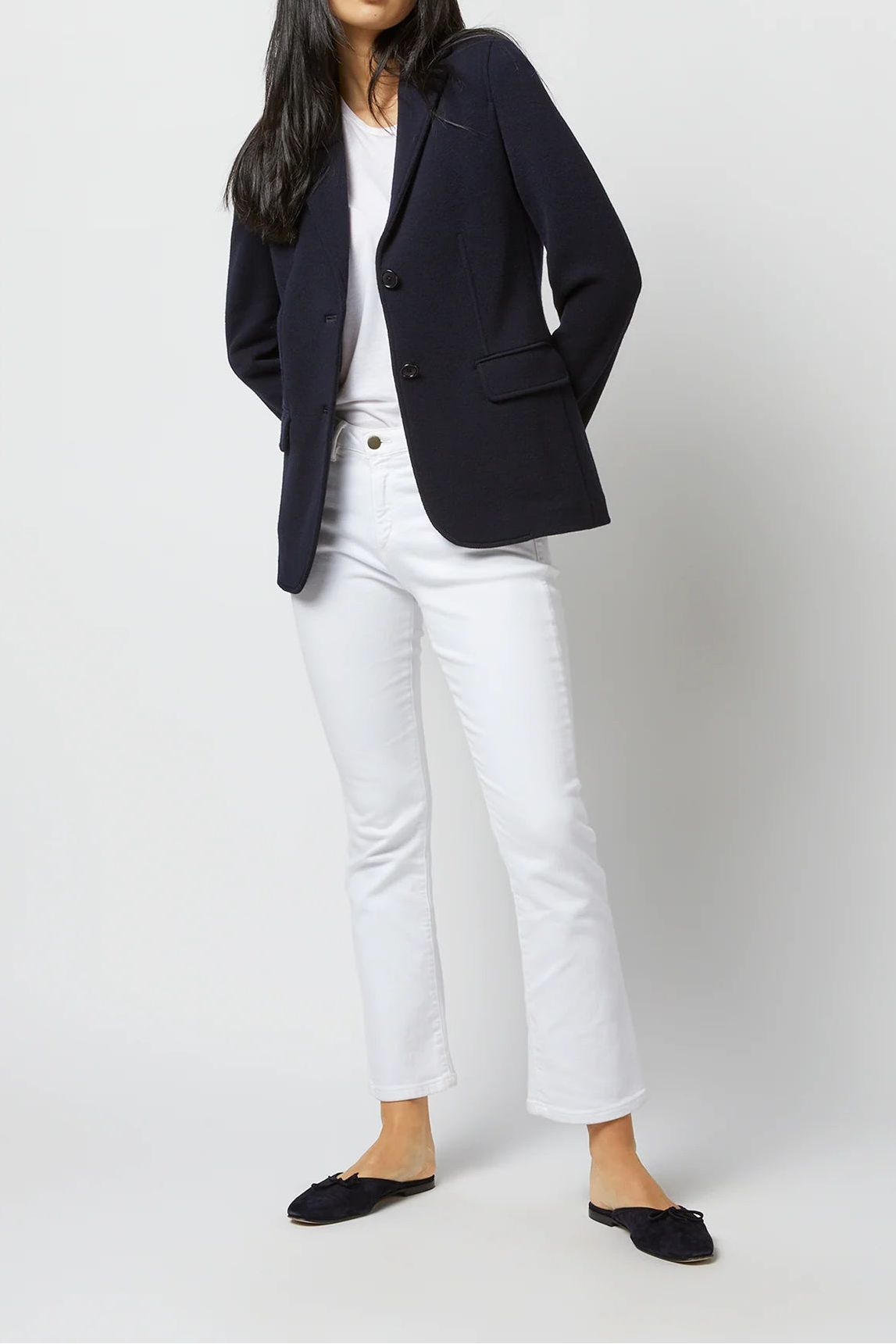 A woman modeling a dark navy blazer over a white top and white cropped jeans, paired with black flats, against a light gray studio background.