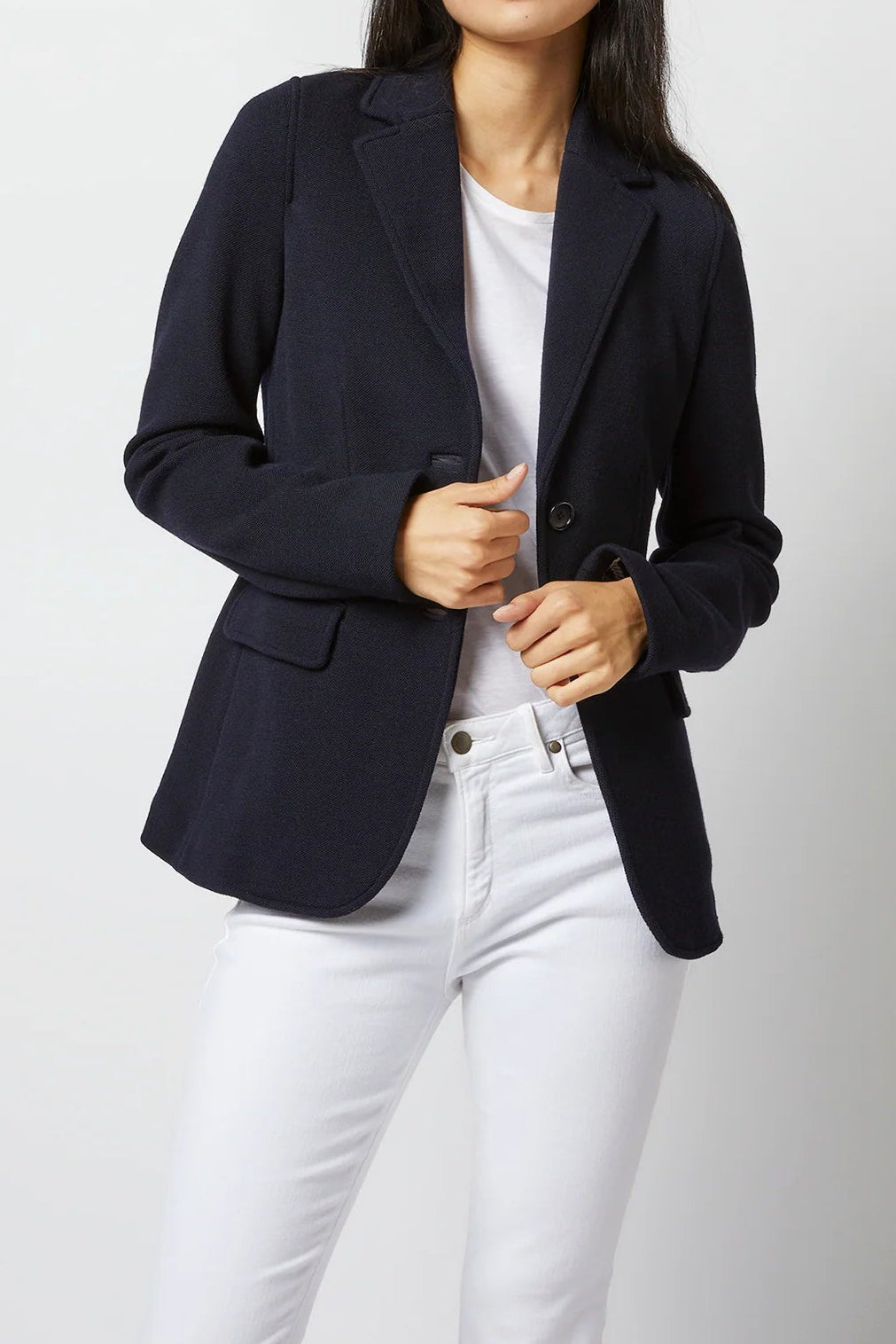 A woman modeling a dark navy blue blazer over a white t-shirt and white jeans, standing against a light gray studio background.