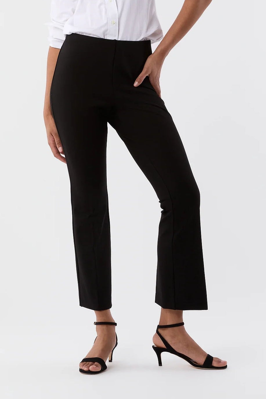 A model wearing a white button-down shirt, black cropped trousers, and black strappy heels, standing against a plain white studio background.