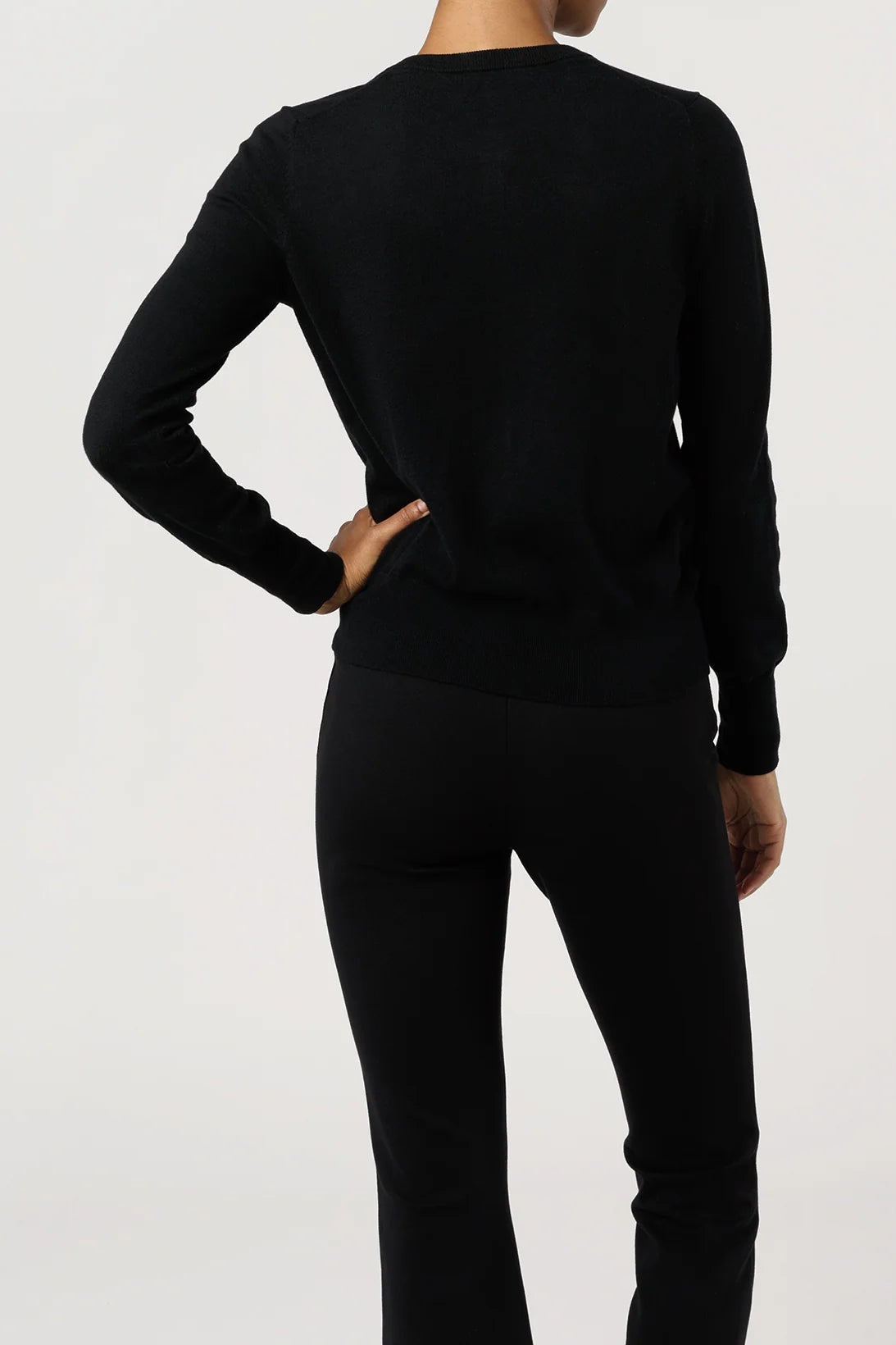 A woman shown from the back wearing a black long-sleeve crewneck top and black pants against a plain light gray studio background.