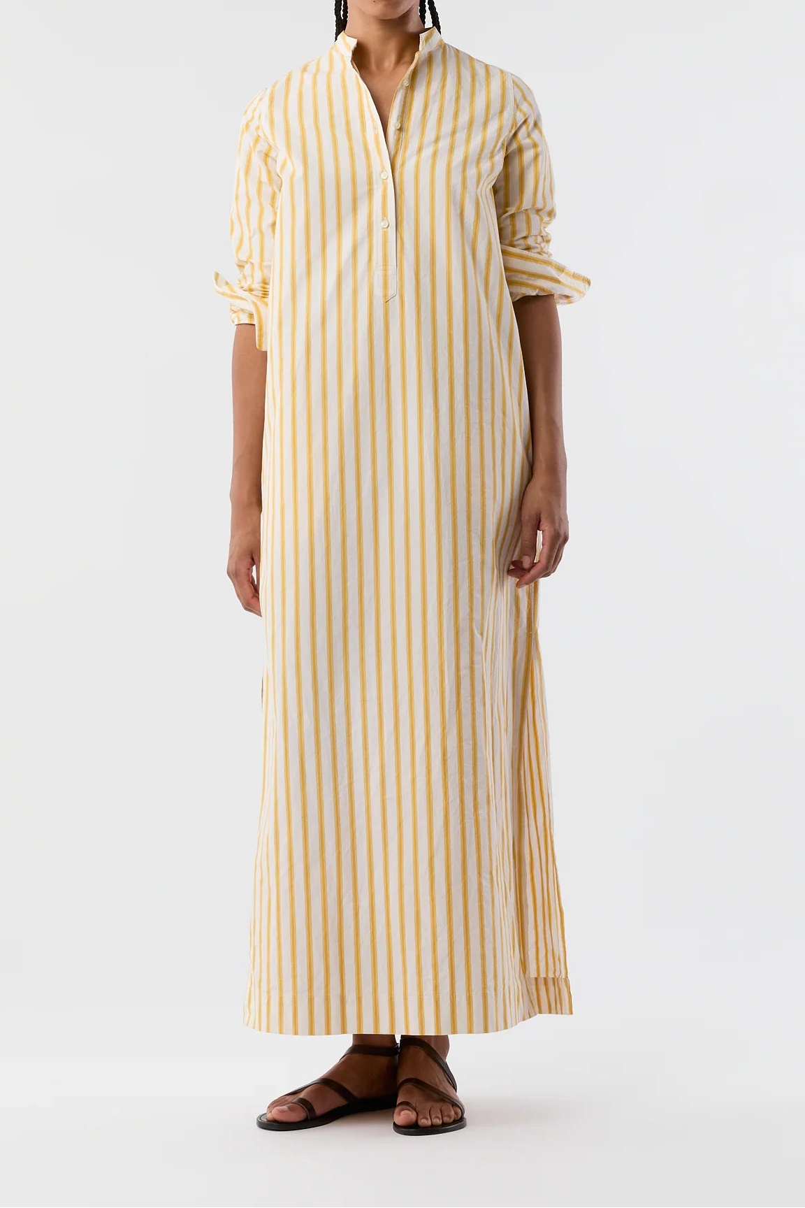 A woman modeling a long-sleeved, collared maxi dress with vertical yellow stripes on a cream background, paired with brown leather sandals, against a plain white studio background.