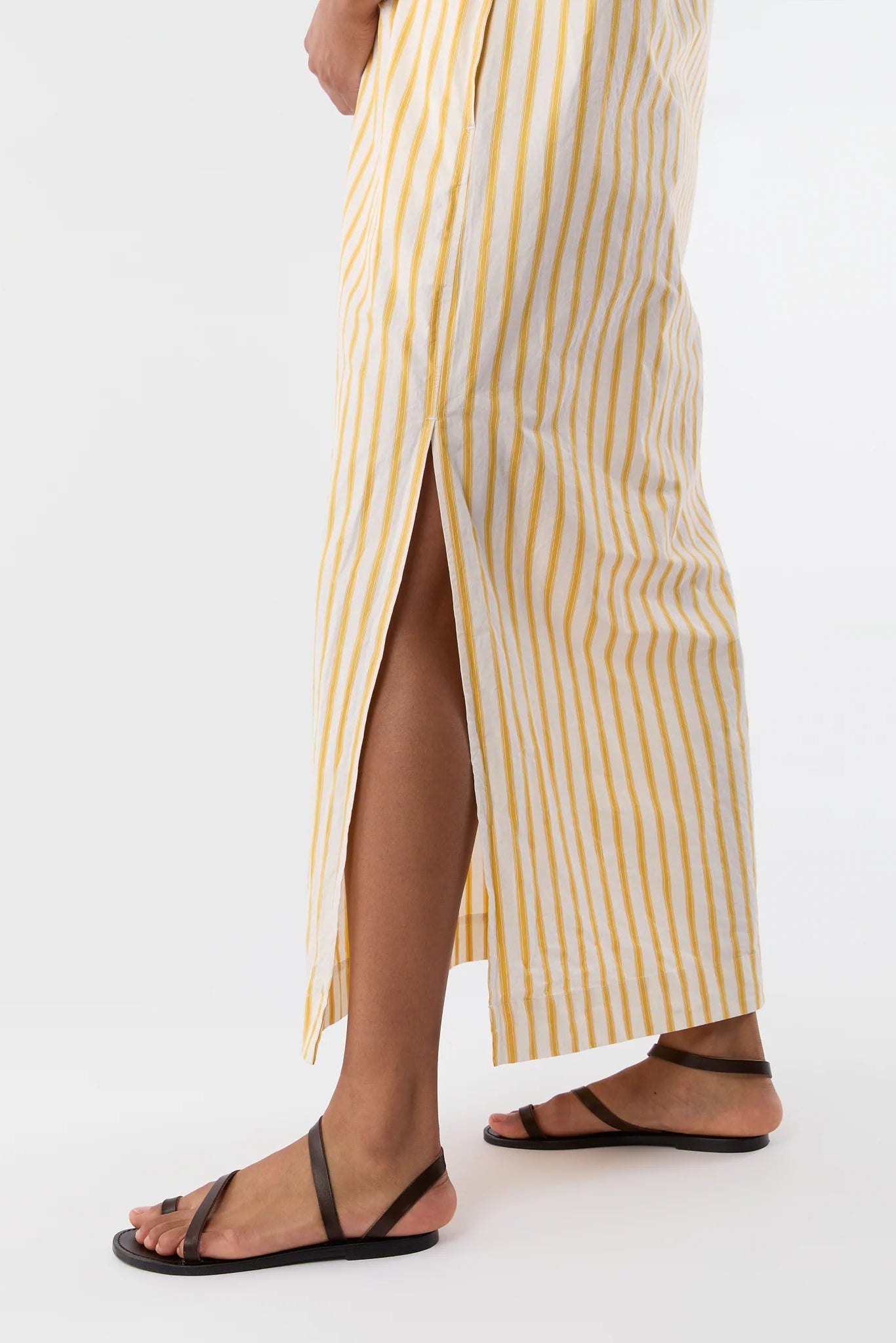 A person wearing a cream and yellow vertically striped wrap skirt with a high side slit, paired with black flat sandals, shown from the waist down against a plain white studio background.