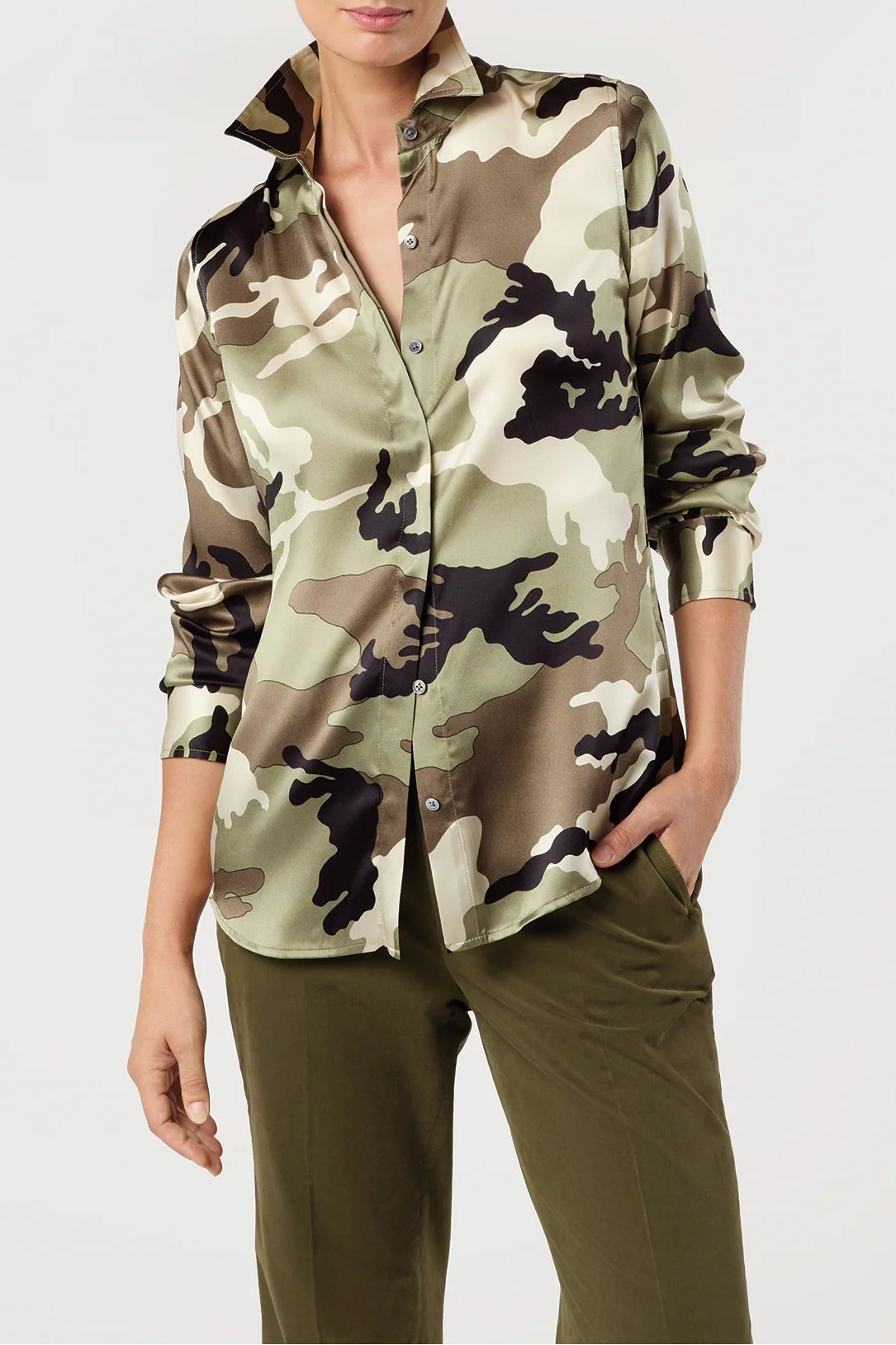 A woman modeling a long-sleeve button-up shirt with a green, beige, and black camouflage pattern, paired with olive green pants, against a plain light gray background.