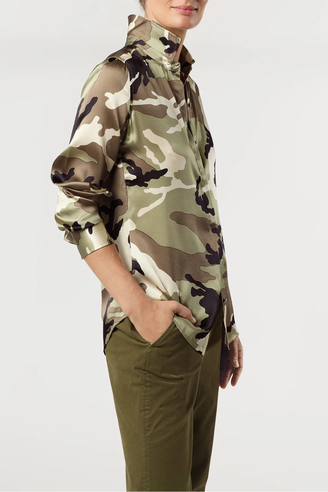 A woman modeling a shiny camouflage print jacket with a high collar and half-zip front, paired with olive green pants, against a plain light gray background.