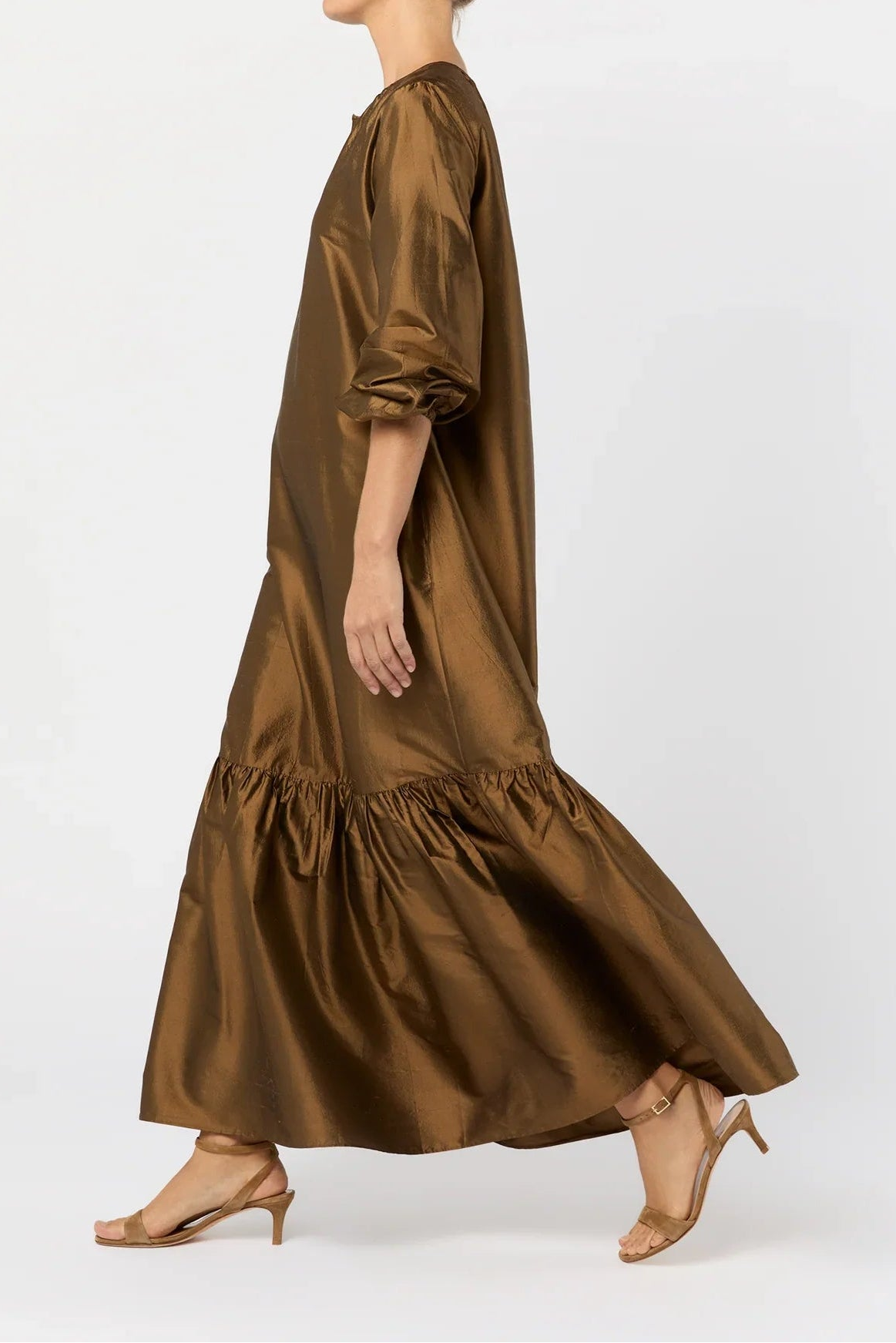 A woman in profile wearing a flowing bronze-brown satin maxi dress with voluminous elbow-length puff sleeves and a gathered tiered hem, paired with tan heeled sandals, against a plain light gray studio background.