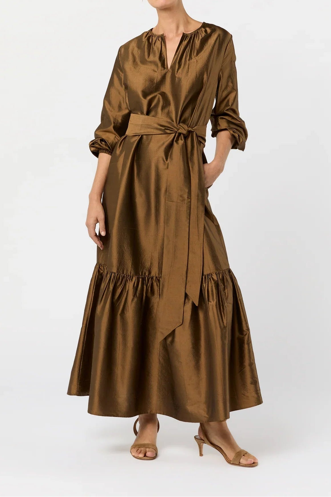 A woman modeling a brown satin midi dress with a tie waist, ruffled sleeves, and a tiered skirt, paired with brown heeled sandals against a light gray studio background.
