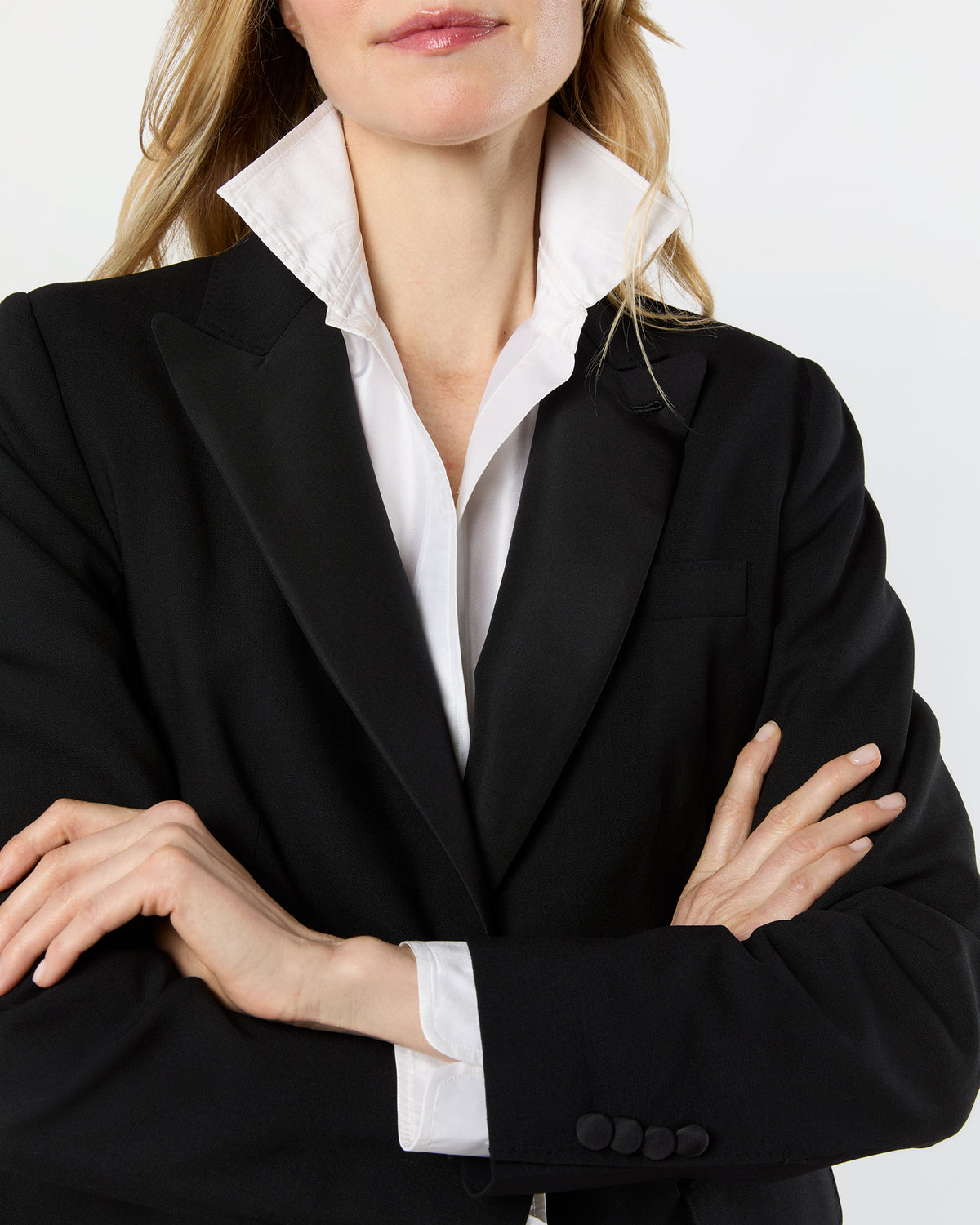 A close-up of a woman wearing a black tailored blazer over a white collared shirt, with her arms crossed against a plain light background.